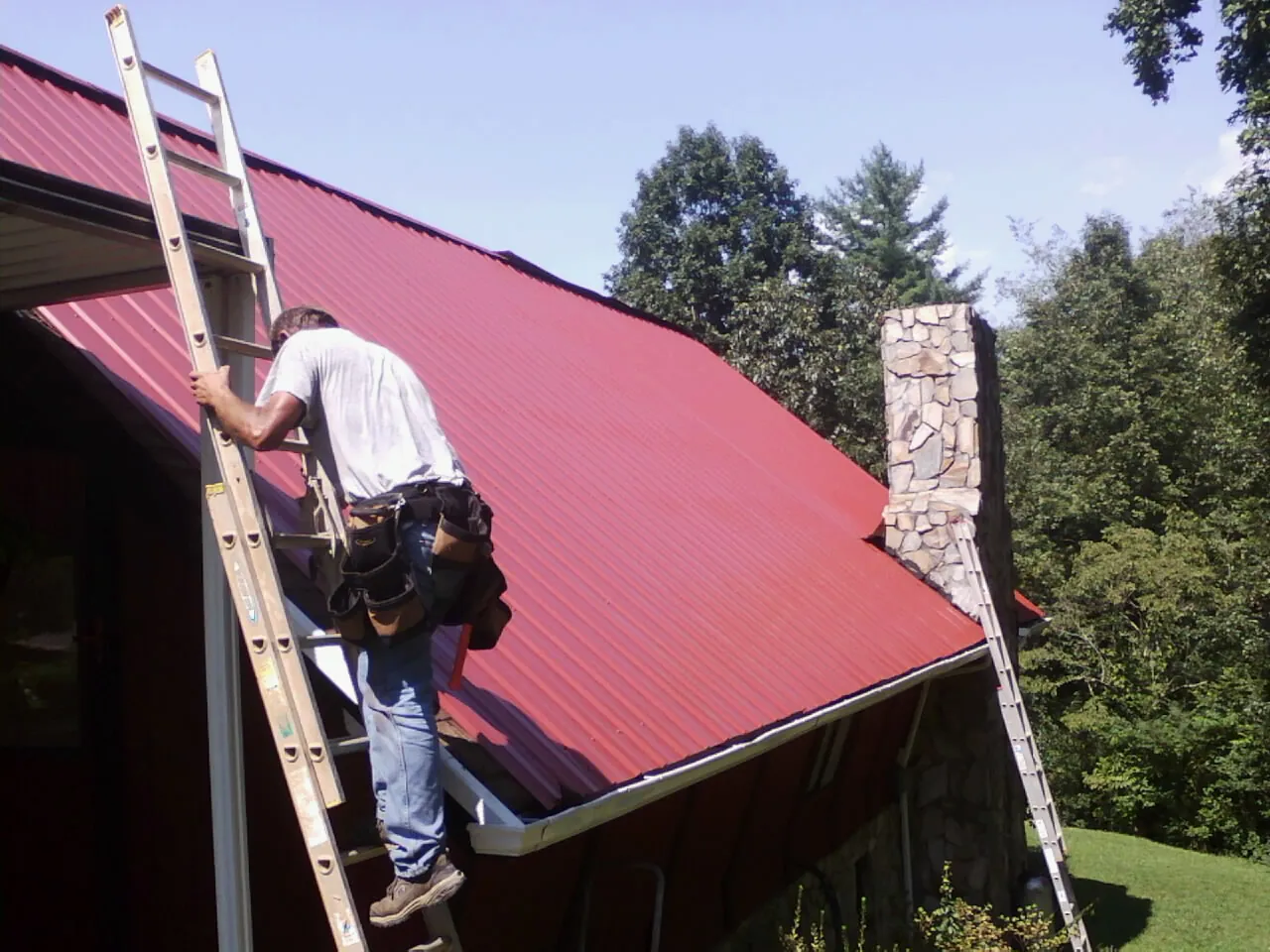 Roofer on a ladder beside a red metal roof and stone chimney — Soffit & Fascia Repair by Roofing Brothers in Vintondale